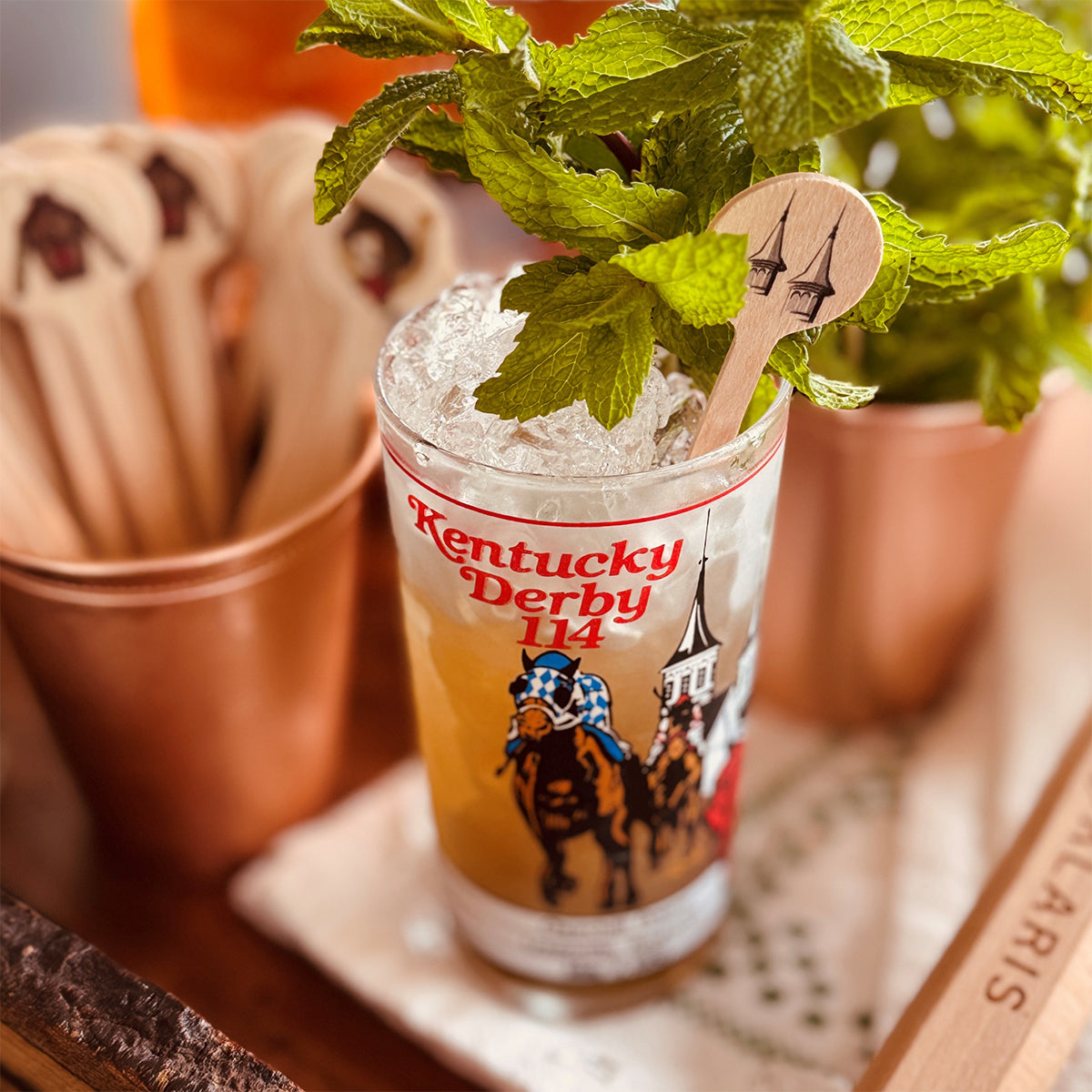 Mint Julep in a glass with mint leaves and a wooden spoon, featuring Kentucky Derby branding.